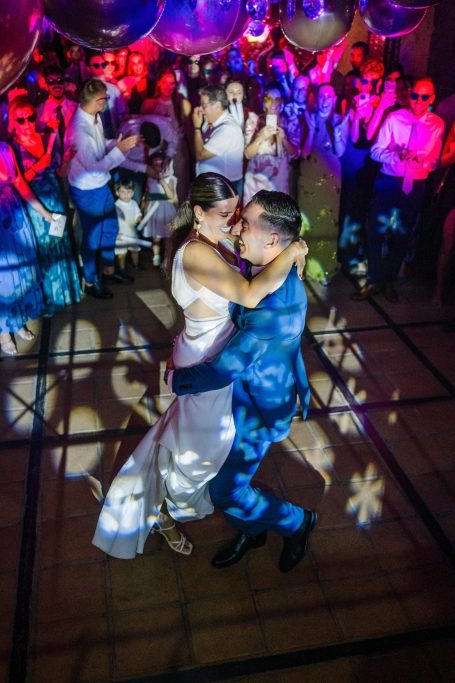 Novios de pelicula Pareja bailando en una pista iluminada, rodeada de invitados en un ambiente festivo.